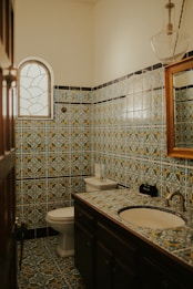 A bathroom with intricate, multicolored patterned tiles covering the walls and floor. The room features a sink with a wooden-framed mirror above it and a round window with a decorative pattern.