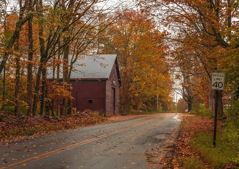 A wet country road lined with orange autumn trees passing an old red barn, typical of the back roads outside Goffstown, NH