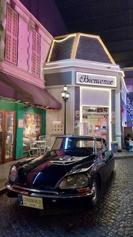 A vintage Citro&euml;n DS 21 car is parked on a cobblestone street in front of a French-themed caf&eacute;. The entrance to the caf&eacute; features a sign that reads 'Bienvenue' and the fa&ccedil;ade is styled with classic architecture. The scene is illuminated with decorative lights and a street lamp.
