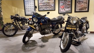 Three vintage motorcycles are displayed on a tiled floor within a room, with framed pictures and posters of cars and motorcycles adorning the yellow walls. The motorcycles have classic designs with visible brand logos and vintage number plates.