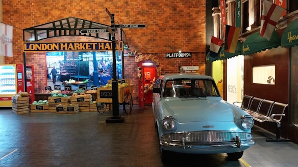A vintage market scene featuring a light blue classic car parked beside a traditional marketplace. The marketplace showcases various crates filled with fruits and vegetables, displayed next to a brick wall with signs indicating it's the 'London Market Hall.' Brightly colored flags and signs, including directions to famous locations like 'Baker Street' and 'Camp Road,' add to the nostalgic ambiance. A red phone booth is visible, enhancing the classic British setting.