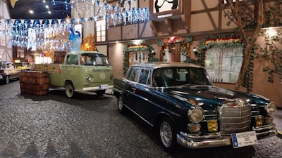 A vintage display featuring a classic green Volkswagen van with barrels on its flatbed parked next to a dark-colored Mercedes Benz. The scene is set on a cobblestone street with festive decorations, including strings of lights and garlands, creating a quaint and nostalgic atmosphere. Surrounding buildings have traditional architecture with wooden beams and floral decorations.