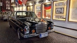 A classic Mercedes Benz car in a museum-like setting with historical images and flags displayed on the wall behind. The floor is cobblestoned, adding a vintage ambiance to the scene.