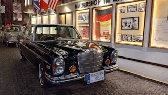 A classic Mercedes Benz car in a museum-like setting with historical images and flags displayed on the wall behind. The floor is cobblestoned, adding a vintage ambiance to the scene.