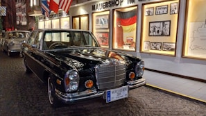 A classic Mercedes Benz car in a museum-like setting with historical images and flags displayed on the wall behind. The floor is cobblestoned, adding a vintage ambiance to the scene.