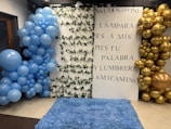 A decorative setup featuring a white backdrop with Spanish text from Psalm 119:50. The backdrop has two large balloon arrangements on each side: blue balloons on the left and gold balloons on the right. The white panel is adorned with intertwined green leaves. The floor is covered with a fluffy blue rug.