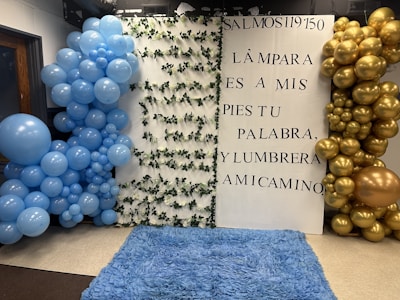 A decorative setup featuring a white backdrop with Spanish text from Psalm 119:50. The backdrop has two large balloon arrangements on each side: blue balloons on the left and gold balloons on the right. The white panel is adorned with intertwined green leaves. The floor is covered with a fluffy blue rug.