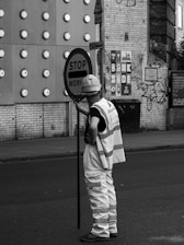 A person wearing a hard hat and reflective vest is holding a sign that reads 'STOP WOR...'. The background features a brick wall with circular protrusions, signage, and some graffiti.