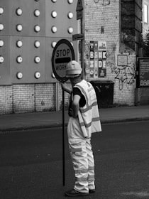 A person wearing a hard hat and reflective vest is holding a sign that reads 'STOP WOR...'. The background features a brick wall with circular protrusions, signage, and some graffiti.