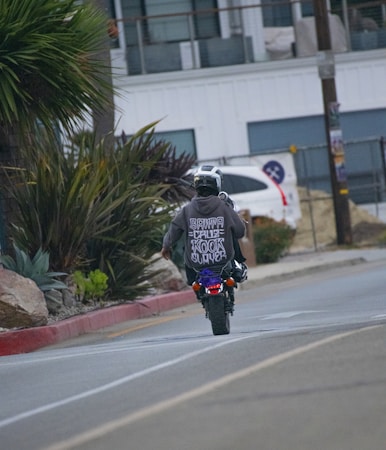 A person wearing a helmet and a dark hoodie is riding a small motorcycle on a street. The back of the hoodie has a bold graphic design. The street is lined with plants and a parked car is visible in the background. The scene appears to be urban with a modern building and some construction materials nearby.