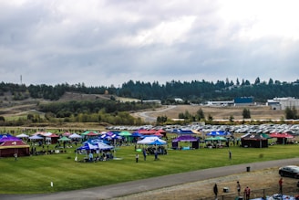 A large outdoor field with multiple colorful tents set up, likely for a festival or event. Numerous people are gathered around the tents. Cars are parked in rows behind the event area. The background features a forested landscape with a cloudy sky overhead.