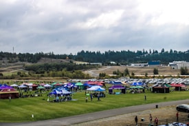 A large outdoor field with multiple colorful tents set up, likely for a festival or event. Numerous people are gathered around the tents. Cars are parked in rows behind the event area. The background features a forested landscape with a cloudy sky overhead.