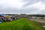 A grassy field with several tents set up along one side, displaying banners with names. People are gathered around the tents, suggesting a community or school event. In the background, there are parked cars and a hill with a line of trees on top, under an overcast sky.
