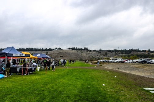 A grassy field with several tents set up along one side, displaying banners with names. People are gathered around the tents, suggesting a community or school event. In the background, there are parked cars and a hill with a line of trees on top, under an overcast sky.