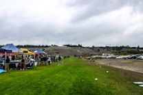 A grassy field with several tents set up along one side, displaying banners with names. People are gathered around the tents, suggesting a community or school event. In the background, there are parked cars and a hill with a line of trees on top, under an overcast sky.
