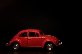 A vintage red car in a model size is positioned against a dark background. The vehicle's design is reminiscent of classic cars with smooth curves and chrome details, particularly visible on the wheels and bumpers. The car's windows are tinted, adding a slight reflection from the surrounding light.