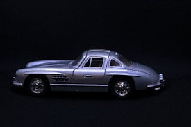 A silver miniature car model, positioned on a dark background, shown in a side profile. The car features a sleek design with detailed body lines, side mirrors, and shiny wheels.
