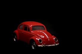 A classic red vintage car model is set against a stark black background. The car has a smooth, rounded design with distinctive headlights and chrome accents.