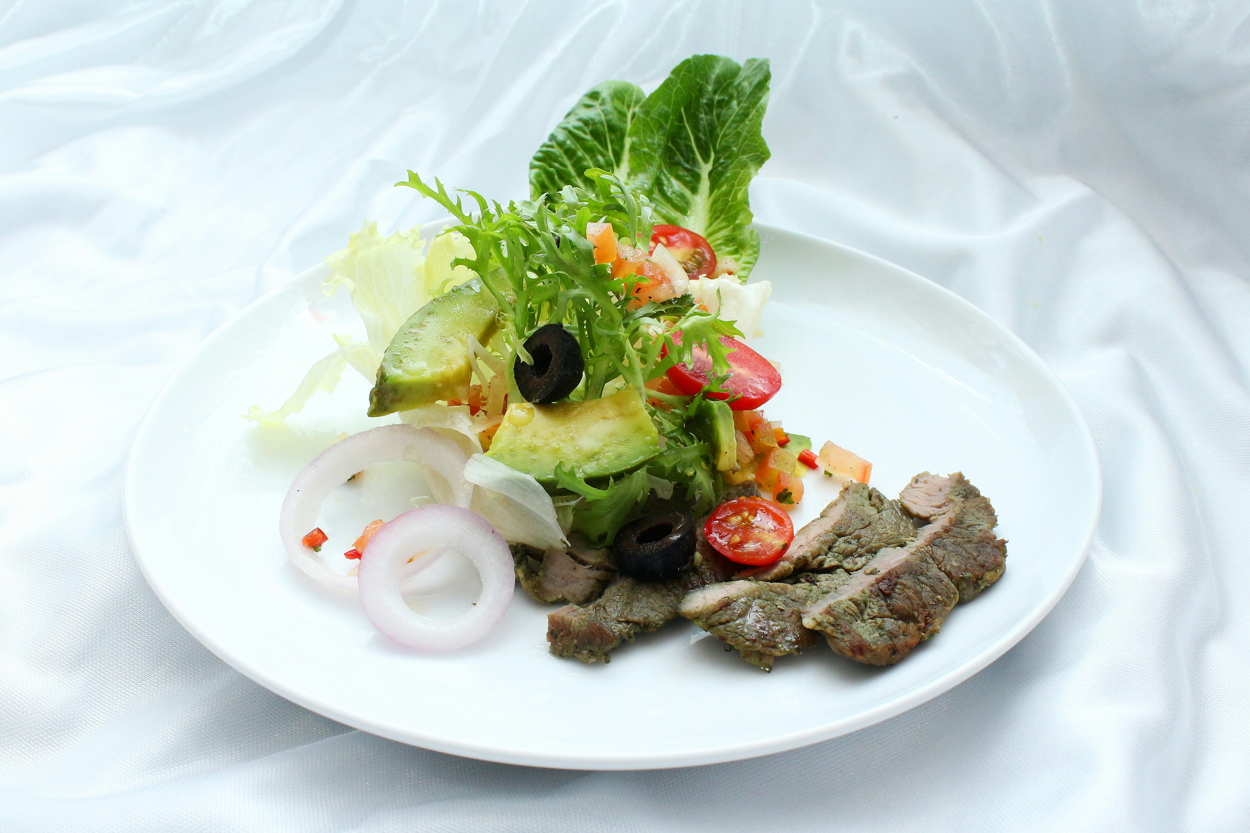 a white plate topped with meat and veggies, Seared beef salad with sliced avocado, tomato, and fresh vegetables. Thinly sliced sashimi style.