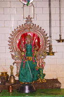 A richly adorned statue is placed against a white tiled wall. It features a figure draped in green fabric with gold accents and decorated with pink flowers. The statue is set within an ornate frame with intricate designs. Surrounding the statue are various metal vessels and traditional hanging lamps.