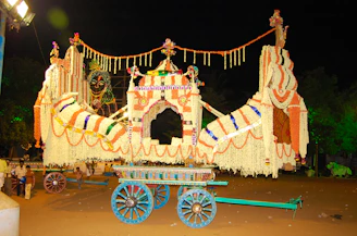 A large, elaborately decorated chariot adorned with colorful garlands and floral designs is illuminated against the night sky. The chariot is ornately embellished with flowers in shades of orange, white, and pink, accompanied by intricate lights depicting a deity. Several people are gathered around, possibly indicating the preparation for or conclusion of a festive event.