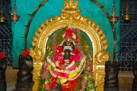 A deity statue adorned with colorful flower garlands and surrounded by a golden ornate arch. There are decorative lamps hanging on either side, and the background features a teal-colored wall with a lattice pattern.