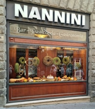A bakery shop window displaying a variety of baked goods and decorative items. The display includes woven wreaths, baskets, and an assortment of breads and pastries. The shop's signage is visible above the window with bold lettering.