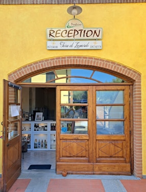 A charming reception entrance with a wooden door and a sign above that reads 'RECEPTION' along with business names and phone numbers. The facade features yellow walls with brick accents. Inside, there is a small reception area displaying various bottles and items on white shelves under soft lighting.