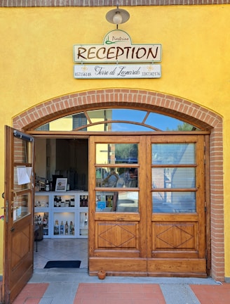 A charming reception entrance with a wooden door and a sign above that reads 'RECEPTION' along with business names and phone numbers. The facade features yellow walls with brick accents. Inside, there is a small reception area displaying various bottles and items on white shelves under soft lighting.
