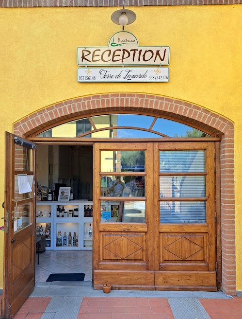 A charming reception entrance with a wooden door and a sign above that reads 'RECEPTION' along with business names and phone numbers. The facade features yellow walls with brick accents. Inside, there is a small reception area displaying various bottles and items on white shelves under soft lighting.
