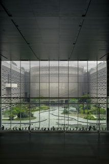 A large, modern architectural interior with a wide glass window overlooking a courtyard. The courtyard features a geometric pathway surrounded by greenery, including various plants and trees. The window structure is made of grid-like metal framing, and the exterior walls have textured patterns.