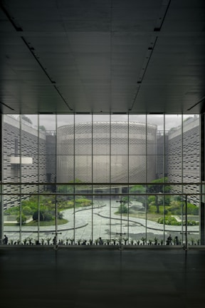 A large, modern architectural interior with a wide glass window overlooking a courtyard. The courtyard features a geometric pathway surrounded by greenery, including various plants and trees. The window structure is made of grid-like metal framing, and the exterior walls have textured patterns.