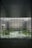 A large, modern architectural interior with a wide glass window overlooking a courtyard. The courtyard features a geometric pathway surrounded by greenery, including various plants and trees. The window structure is made of grid-like metal framing, and the exterior walls have textured patterns.
