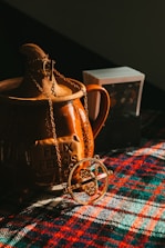 A golden necklace with a circular pendant is draped over a brown ceramic container with the inscription 'Felix Felicis'. The scene is set on a colorful plaid fabric, and there is a small stack of cards in the background.
