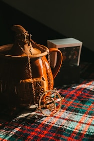 A golden necklace with a circular pendant is draped over a brown ceramic container with the inscription 'Felix Felicis'. The scene is set on a colorful plaid fabric, and there is a small stack of cards in the background.