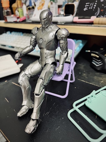 A detailed model of a silver armored figure is seated on a small purple folding chair. The background includes various desk items such as a keyboard, electronic devices, and stationery, indicating a cluttered workspace.