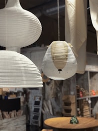 A cozy interior setting featuring multiple hanging paper lanterns, one with a cute animal face design. Shelving units and decorative items are seen in the background alongside a wooden circular table with a small decorative pot.