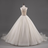 A mannequin displays an elegant white ball gown with a fitted bodice adorned with intricate beadwork and a voluminous layered skirt.