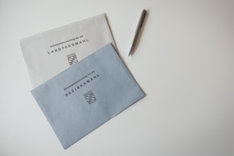 Two election envelopes are lying on a white surface. One envelope is labeled 'Landtagswahl' and the other 'Bezirkswahl,' both featuring a crest symbol. A pen with a metallic finish is placed nearby.