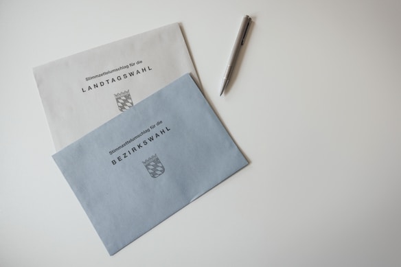 Two election envelopes are lying on a white surface. One envelope is labeled 'Landtagswahl' and the other 'Bezirkswahl,' both featuring a crest symbol. A pen with a metallic finish is placed nearby.