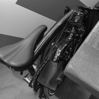 A black and white image of a folded bicycle with curved seat and visible chain, placed near a geometric patterned wall and next to a wooden surface.