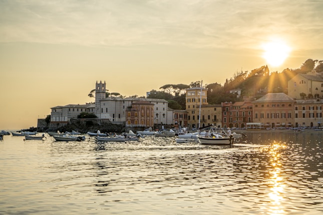 A picturesque coastal townscape bathed in the warm light of a setting sun. Elegant buildings with varied architecture line the waterfront, with small boats gently floating on the calm water, their reflections shimmering in the golden glow.