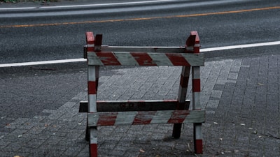 A wooden road barrier painted with alternating red and white diagonal stripes stands on a paved surface. The background features an asphalt road with a visible white line and a yellow line. Leaves and debris are scattered near the barrier, suggesting an outdoor setting.