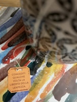 A colorful watercolor background features an assortment of brushstrokes in various hues. In the foreground, a ceramic object with intricate designs is partially visible. A paper tag with Italian text is attached by a string, adding a personal or artistic touch to the composition.