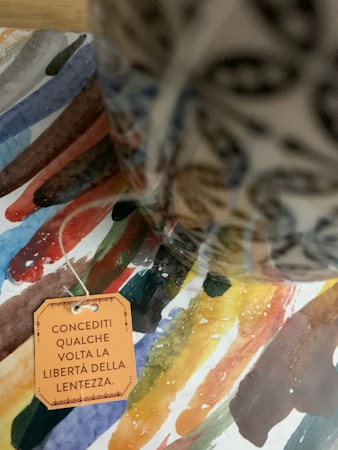 A colorful watercolor background features an assortment of brushstrokes in various hues. In the foreground, a ceramic object with intricate designs is partially visible. A paper tag with Italian text is attached by a string, adding a personal or artistic touch to the composition.