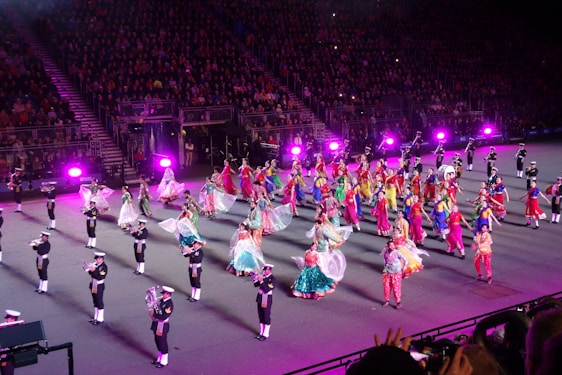 A large group of performers in colorful, traditional costumes are dancing on a stage. The dancers are surrounded by musicians dressed in uniform, playing various brass instruments. The performance is taking place in a stadium filled with a large audience, with bright purple stage lights illuminating the scene.