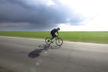 A cyclist in motion is riding a bicycle on an open road. The background features an expansive green field under a cloudy sky, suggesting an outdoor rural setting. The cyclist appears to be dressed in dark attire and is focused on speed, evident by their aerodynamic posture.