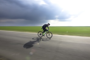 A cyclist in motion is riding a bicycle on an open road. The background features an expansive green field under a cloudy sky, suggesting an outdoor rural setting. The cyclist appears to be dressed in dark attire and is focused on speed, evident by their aerodynamic posture.