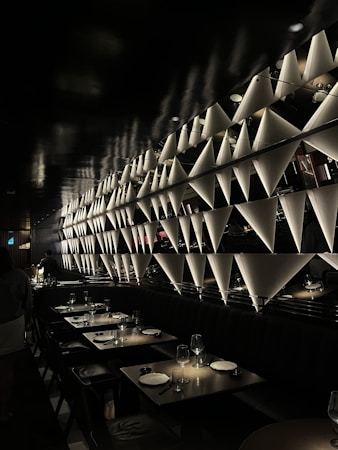 A dimly lit restaurant interior features a uniquely designed wall with triangular light fixtures that cast shadows, creating an artistic ambiance. The seating area comprises dark-colored tables and chairs, with some tables set with plates and glassware. The overall atmosphere appears elegant and modern.