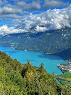 A picturesque landscape featuring a vibrant turquoise lake surrounded by lush green forests. Majestic mountains rise in the background, partially covered by thick, fluffy clouds. A small village is visible along the shoreline, adding charm to the serene natural setting.
